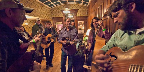 A group of musicians is playing instruments including guitars, a mandolin, and a double bass in a warmly lit, wood-paneled room. They are gathered closely together, fully engaged in their bluegrass performance at the Durango Meltdown, with patterned wallpaper and a wooden floor visible in the background.