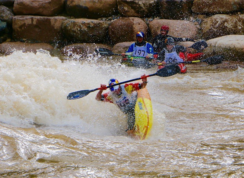 A kayaker in a yellow kayak paddles fiercely through rough whitewater during Animas River Days, while two other kayakers in the background navigate the turbulent river. Large rocks line the riverbank. All kayakers wear helmets and numbered vests, indicating a competitive event.