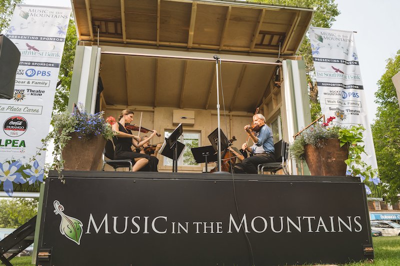 A man and a woman play string instruments on a stage labeled "Music in the Mountains" at the festival. The outdoor stage is surrounded by trees and greenery, with banners displaying sponsor logos on either side. The weather appears clear and sunny, adding to the charming atmosphere.