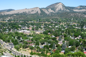 Aerial view of a town nestled in a green landscape with a backdrop of large, rugged mountains under a clear blue sky. The town is surrounded by dense trees and a network of roads.