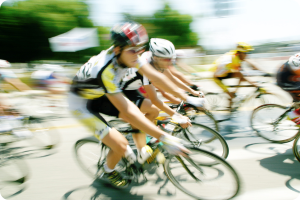 A group of cyclists in bright racing gear speeds through a road during a race. The motion blur emphasizes their fast pace, with trees and a blurry banner in the background on a sunny day.