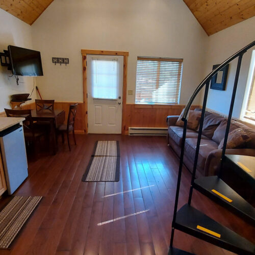 Cozy cabin interior with wood floors, a small kitchenette, dining table, brown couch, wall-mounted TV, spiral staircase, and windows letting in natural light. A door and two rugs are visible on the floor.