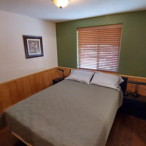 A small bedroom with wood-paneled walls, a green accent wall, a bed with gray bedding, two pillows, a window with wooden blinds, a framed picture, and two black nightstands with lamps.
