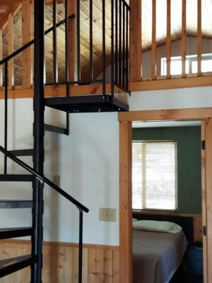 A cozy cabin interior with a black spiral staircase leading to a loft, wood-paneled walls and ceiling, and a bedroom visible through an open doorway. Light streams in from a window by the bed.