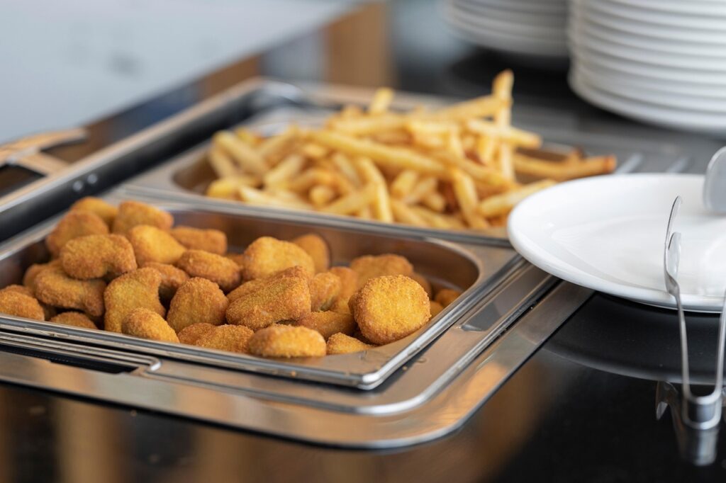 A buffet tray filled with breaded chicken nuggets and another tray with French fries. An empty white plate and serving tongs are placed beside the trays, with stacked plates in the background.