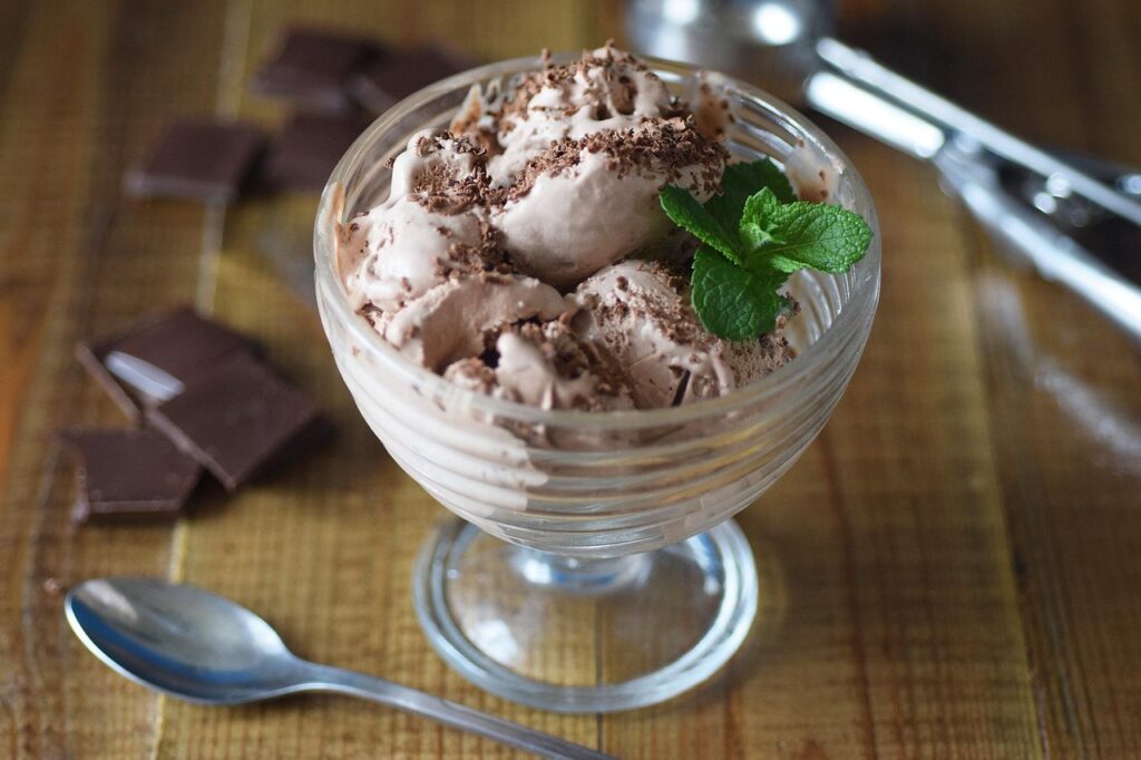 A glass bowl of chocolate ice cream topped with chocolate shavings and a sprig of mint, with a spoon and chocolate pieces on a wooden table.