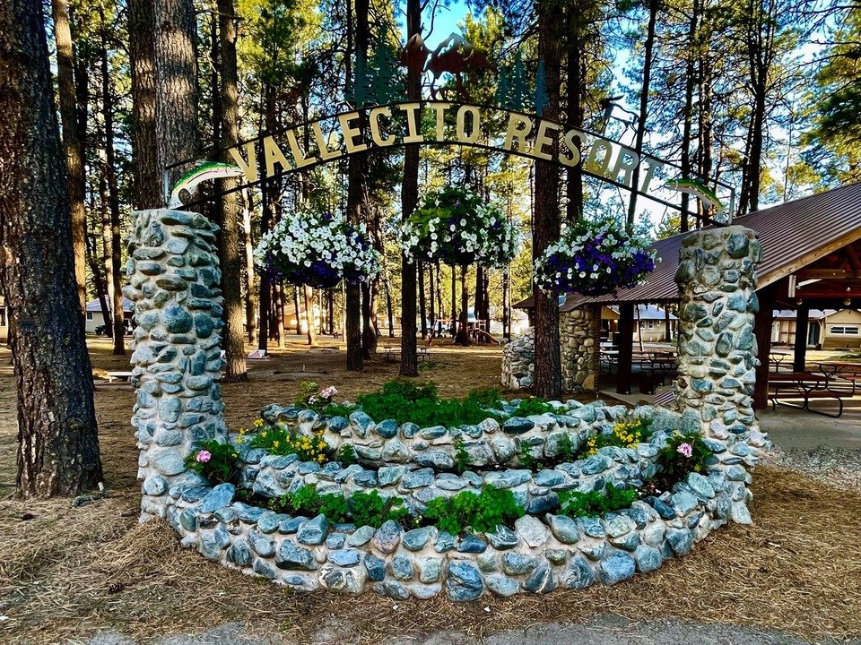 A stone planter bed with flowers sits beneath a sign reading "Vallecito Resort," surrounded by tall pine trees and wooden picnic shelters—one of the scenic JW Locations.