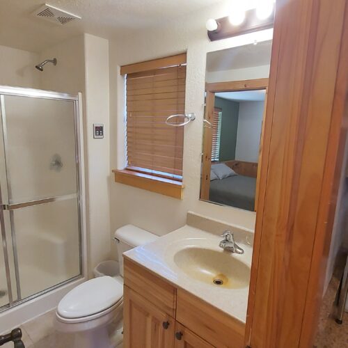 A small bathroom with a shower stall, toilet, and single-sink vanity. There is a window with blinds above the toilet, a mirror above the sink, and wooden accents throughout. Part of a kitchen counter with appliances is visible.