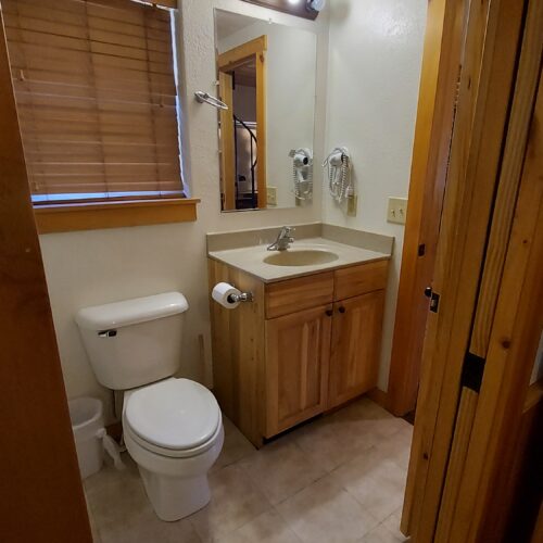 Small bathroom with a closed wooden window blind, toilet, trash can, and wooden vanity with sink, mirror, and wall-mounted lights above. The walls are white, and the floor is tiled. Door is partially open.