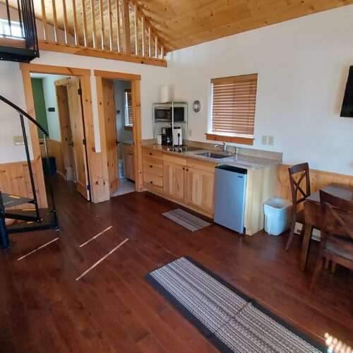 A cozy cabin interior with wood floors and walls, a small kitchen, dining table with chairs, wall-mounted TV, spiral staircase leading to a loft, and natural light from a window with blinds.