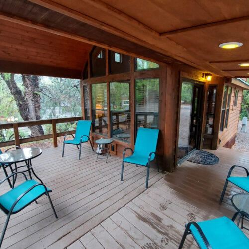 A covered wooden porch with blue chairs and glass tables overlooks a wooded area. Large windows offer a glimpse inside a cozy cabin. The porch has overhead lighting and a mat by the entrance door.