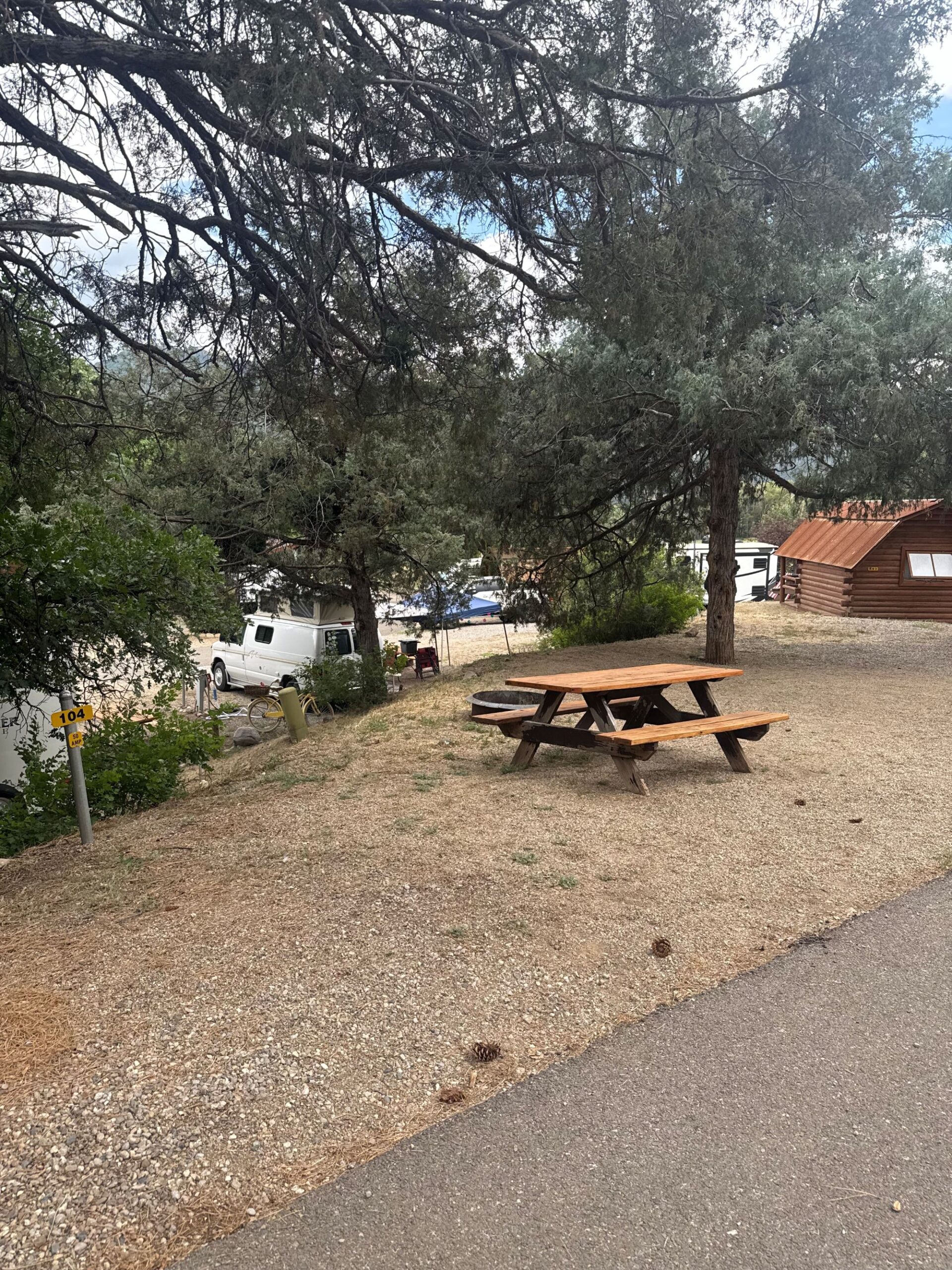 A wooden picnic table sits on a gravel and dirt area under trees near a paved path, with parked vehicles and a small cabin visible in the background.