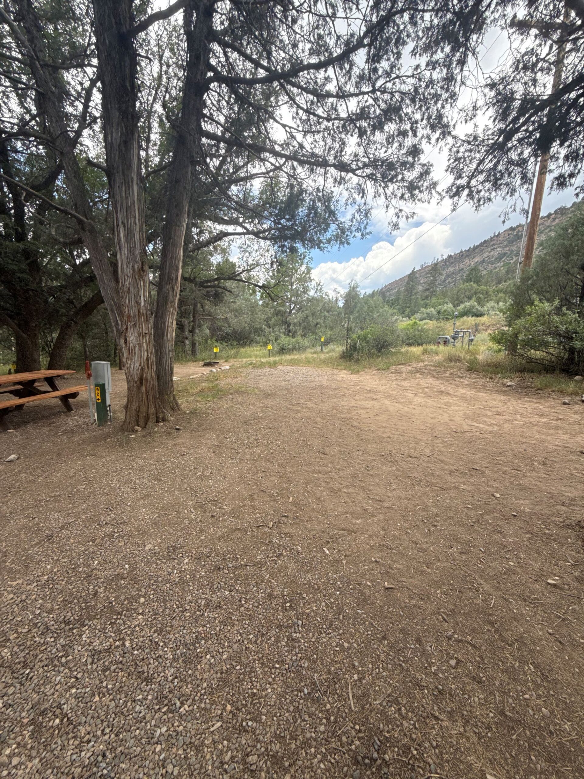 A dirt and gravel clearing with a picnic table on the left, surrounded by trees and hills under a cloudy sky.