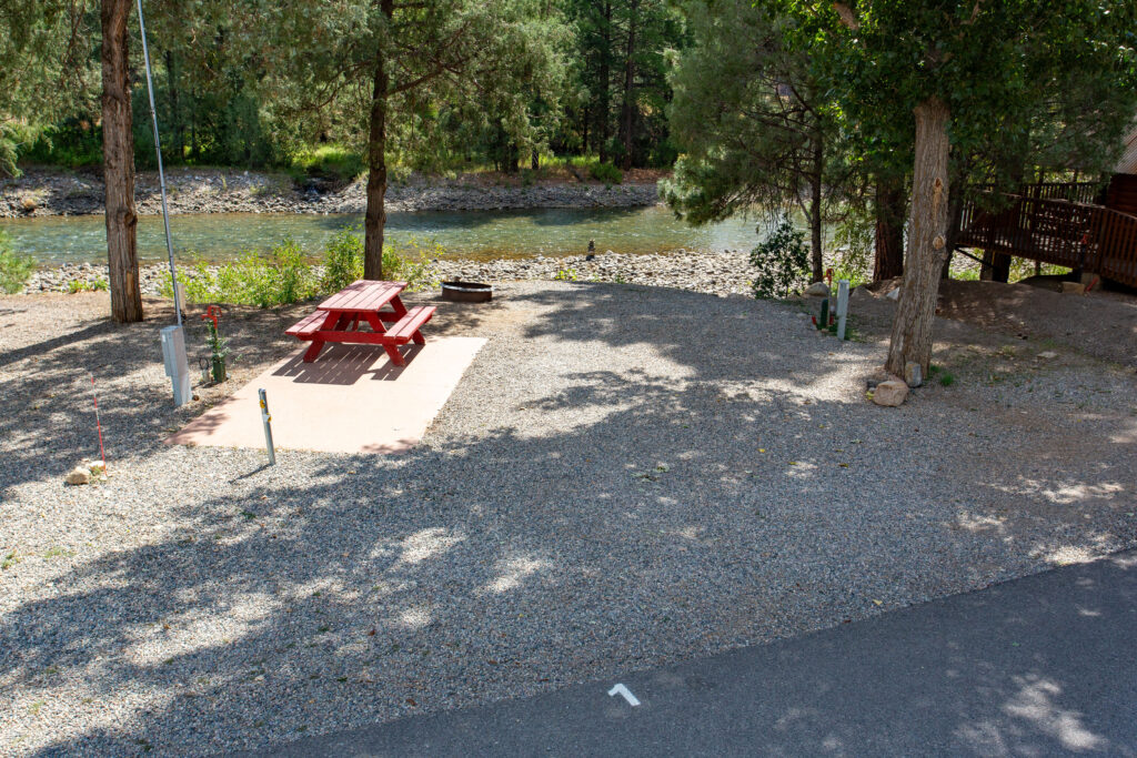 A gravel campsite with a red picnic table and fire ring by a river, surrounded by trees, and electrical hookups visible.