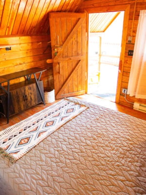A cozy wooden bedroom with a quilted bed, small fridge, woven baskets, a table, and an open door letting in sunlight. White curtains cover a window on the right.