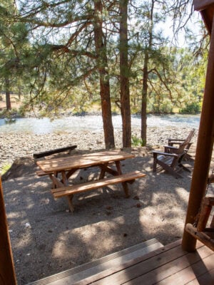 View from a wooden cabin porch overlooking a picnic table, Adirondack chairs, trees, and a riverbank in a natural setting.