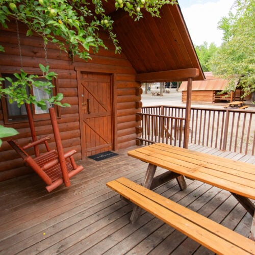 A wooden porch with a picnic table, benches, and a swinging chair outside a log cabin, surrounded by trees.