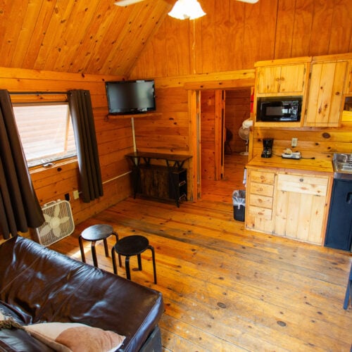 Interior of a small wooden cabin with a leather couch, two stools, a TV, kitchenette, and a dining table, featuring wood-paneled walls, floors, and ceiling.