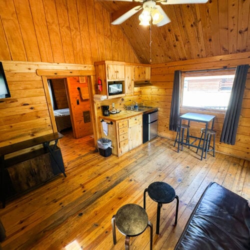A cozy wooden cabin interior with a small kitchen, bar table, stools, TV, and a door leading to a bedroom. Natural light enters through a window with dark curtains.
