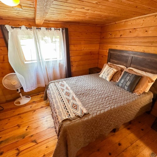Small bedroom with wood-paneled walls and floor, a double bed with patterned bedding, a fan by the window, and a nightstand. Natural light enters through a curtained window.