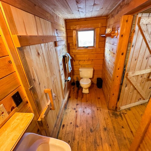 A rustic wooden bathroom with a toilet, sink, towel rack, small window, and wooden door; the walls, floor, and ceiling are all made of wood panels.