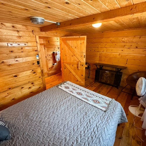 Small wooden bedroom with a quilted bed, a patterned blanket, a fan, a chair, a black heater or fireplace, and a sliding barn door letting in natural light through a window.