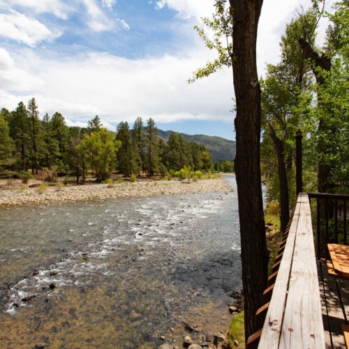 A clear river flows past a forested area under a partly cloudy sky, with a wooden deck and picnic table visible on the right side.