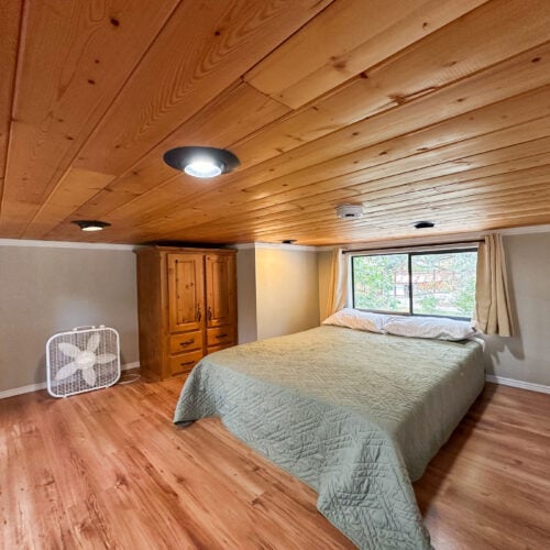 Bedroom with wood-paneled ceiling and floors, a bed with a green quilt, a window with curtains, a wooden wardrobe, and a box fan in the corner.