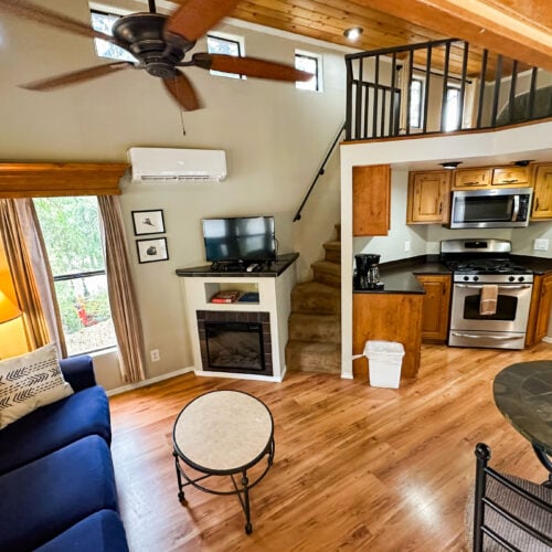 A cozy living area with a blue sofa, round coffee table, fireplace, TV, staircase to a loft, and a kitchen with stainless steel appliances and wooden cabinets.