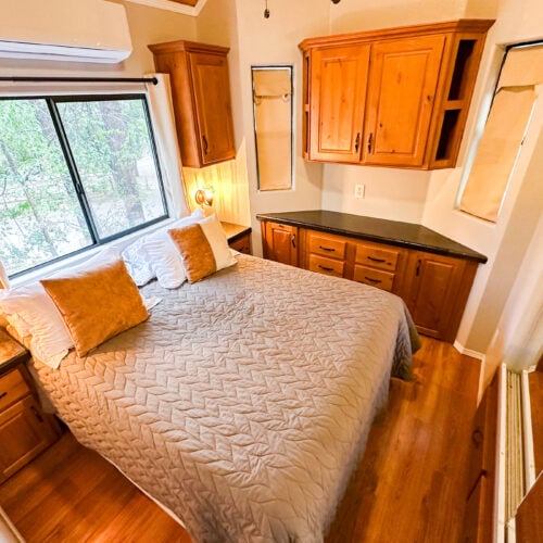 A small bedroom with a double bed, two gold pillows, wooden cabinets, a window with blinds, and hardwood flooring.