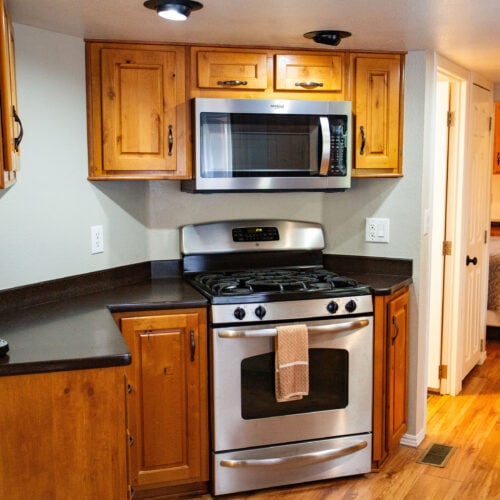 A small kitchen with wooden cabinets, a stainless steel gas stove and microwave, a coffee maker on the counter, and a view into a bedroom with a bed.