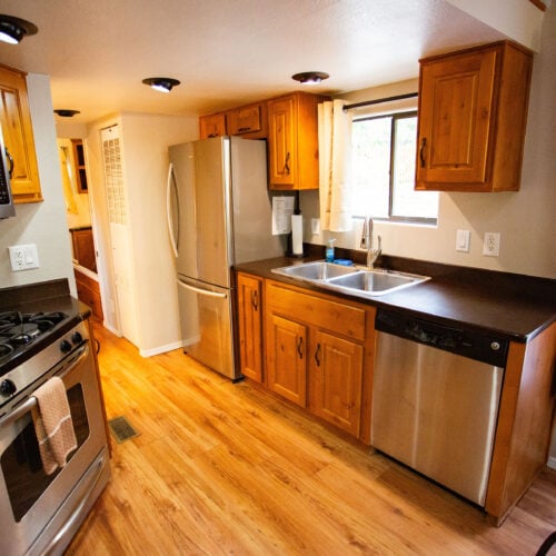 Modern kitchen with wooden cabinets, stainless steel appliances, double sink, and wood flooring, illuminated by natural light from a window.