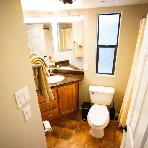 Small bathroom with a brown tiled floor, a white toilet, a sink with a corner countertop, a mirror, and a window. Towels hang near the sink and a shower curtain is visible.