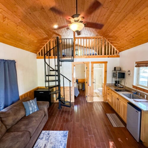 Interior of a small cabin with wood-paneled ceiling and floors, a spiral staircase to a loft, a kitchen area, a brown couch, and a window with blue curtains.