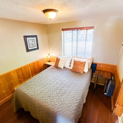 Small bedroom with a double bed, gray quilt, decorative pillows, wooden walls, a window with blinds, bedside table with a lamp, and framed artwork on the wall.