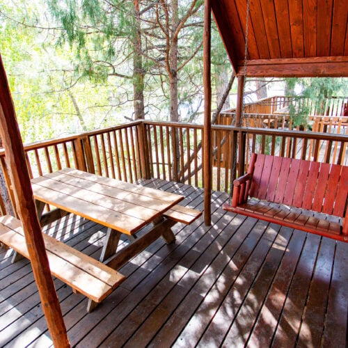 A wooden deck with a picnic table and a red porch swing, surrounded by trees and a wooden railing, attached to a log cabin.