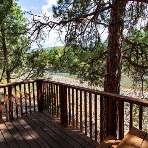 A wooden deck overlooks a rocky riverbank surrounded by tall pine trees and greenery under a partly cloudy sky.