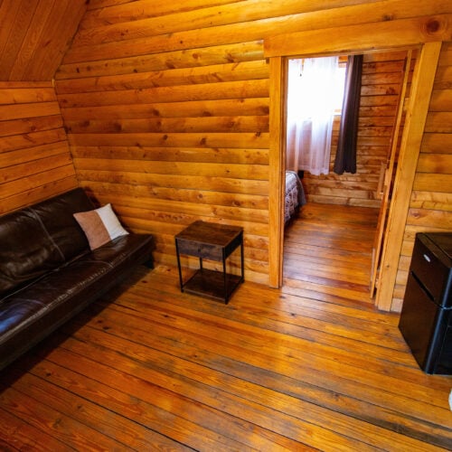 A small wooden cabin interior with a black leather sofa, side table, mini fridge, microwave, and a doorway leading to a bedroom with a curtained window.