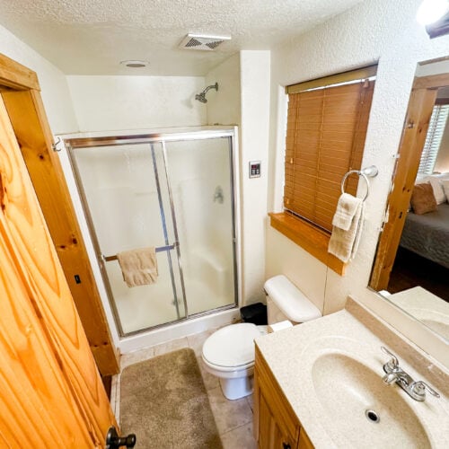 Small bathroom with a shower stall, toilet, sink with countertop, mirror, wooden door, window with blinds, and towel racks; a bedroom is visible through an open doorway.