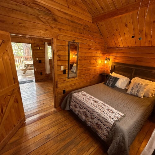 A cozy wooden cabin bedroom with a double bed, patterned bedding, pillows, wall-mounted lamps, a mirror, and an open door leading to a deck with a picnic table.