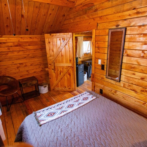 A cozy wooden cabin bedroom with a bed, a patterned runner, a mirror on the wall, a chair, an open door, and natural light coming through windows.