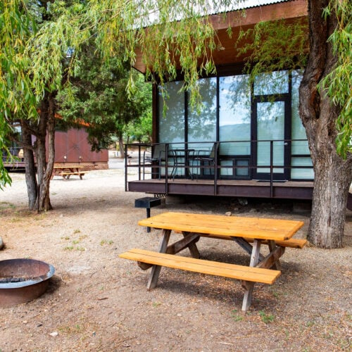 A small cabin with large windows sits between trees, next to a wooden picnic table and a fire ring in a campground setting.