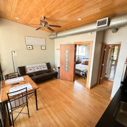 A modern studio apartment with wooden floors, a small dining table, a black sofa, and an open door showing a bedroom with a bed.