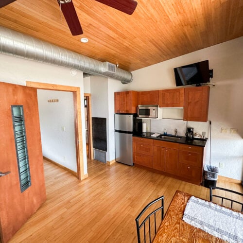 A small modern kitchen with wooden cabinets, stainless steel appliances, a mounted TV, and a dining table, in a room with wood floors and ceiling.