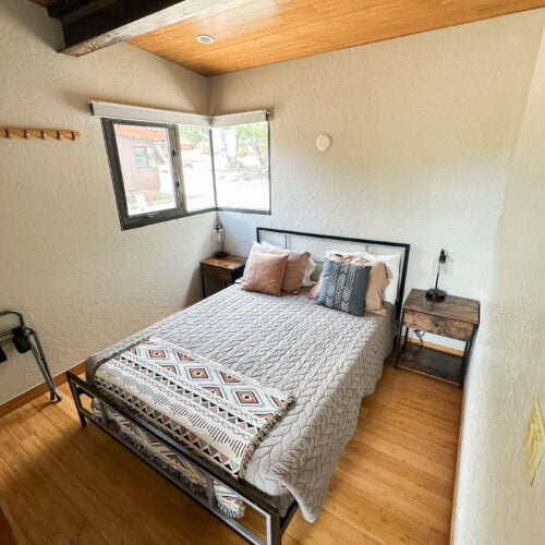 A small bedroom features a neatly made bed with patterned pillows, two wooden nightstands, a window, and wood floors and ceiling.