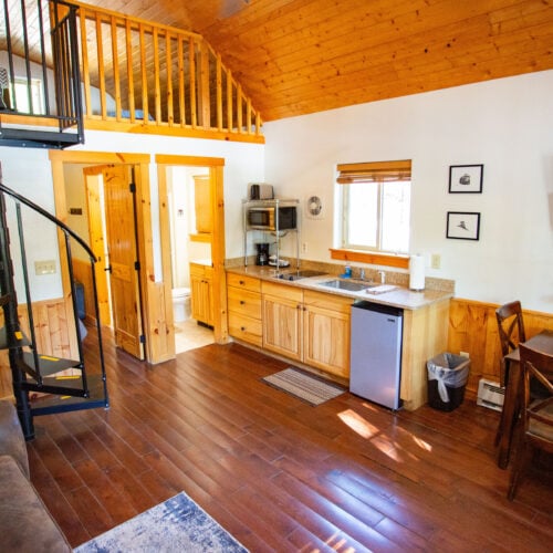 Interior of a small wooden cabin featuring a spiral staircase, kitchenette, dining table, sofa, mounted TV, and a loft sleeping area.