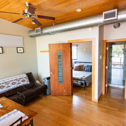 A small, modern apartment with wood flooring, a living area, a visible bedroom, ceiling fan, and exposed air duct.