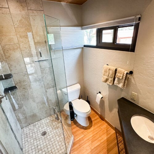 Modern bathroom with a glass-enclosed shower, white toilet, vanity with sink, wall-mounted towels, and wood flooring.