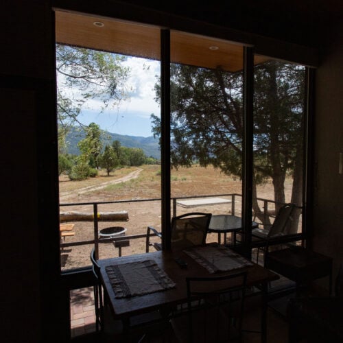 A view through large windows showing a table and chairs inside, with outdoor scenery of trees, a picnic table, and mountains in the background.