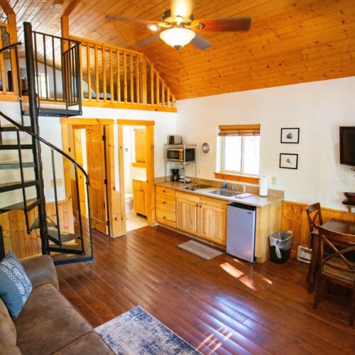 Cozy cabin interior with wood-paneled walls and ceiling, spiral staircase to loft, kitchen area, dining table, wall-mounted TV, and sofa.